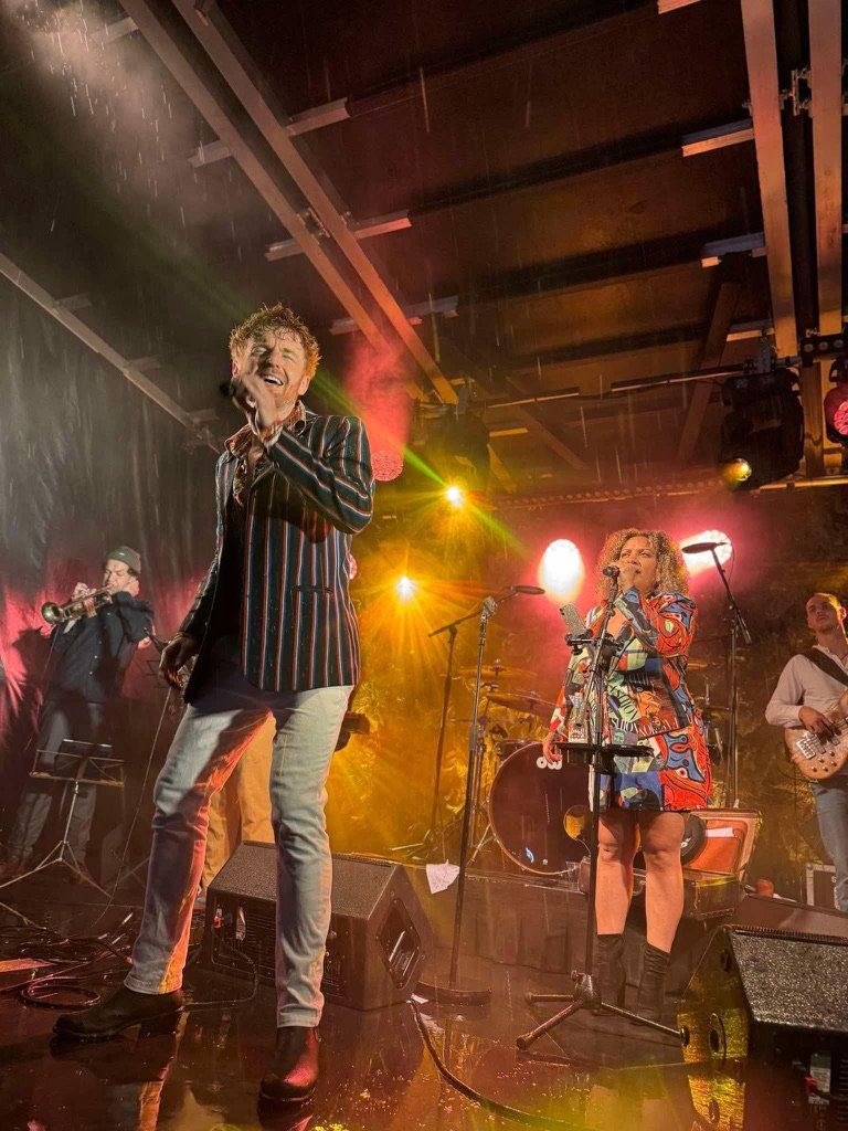 Dillon Rhodes fronting his band mid-set, singing under warm amber stage lights with brass and rhythm section behind
