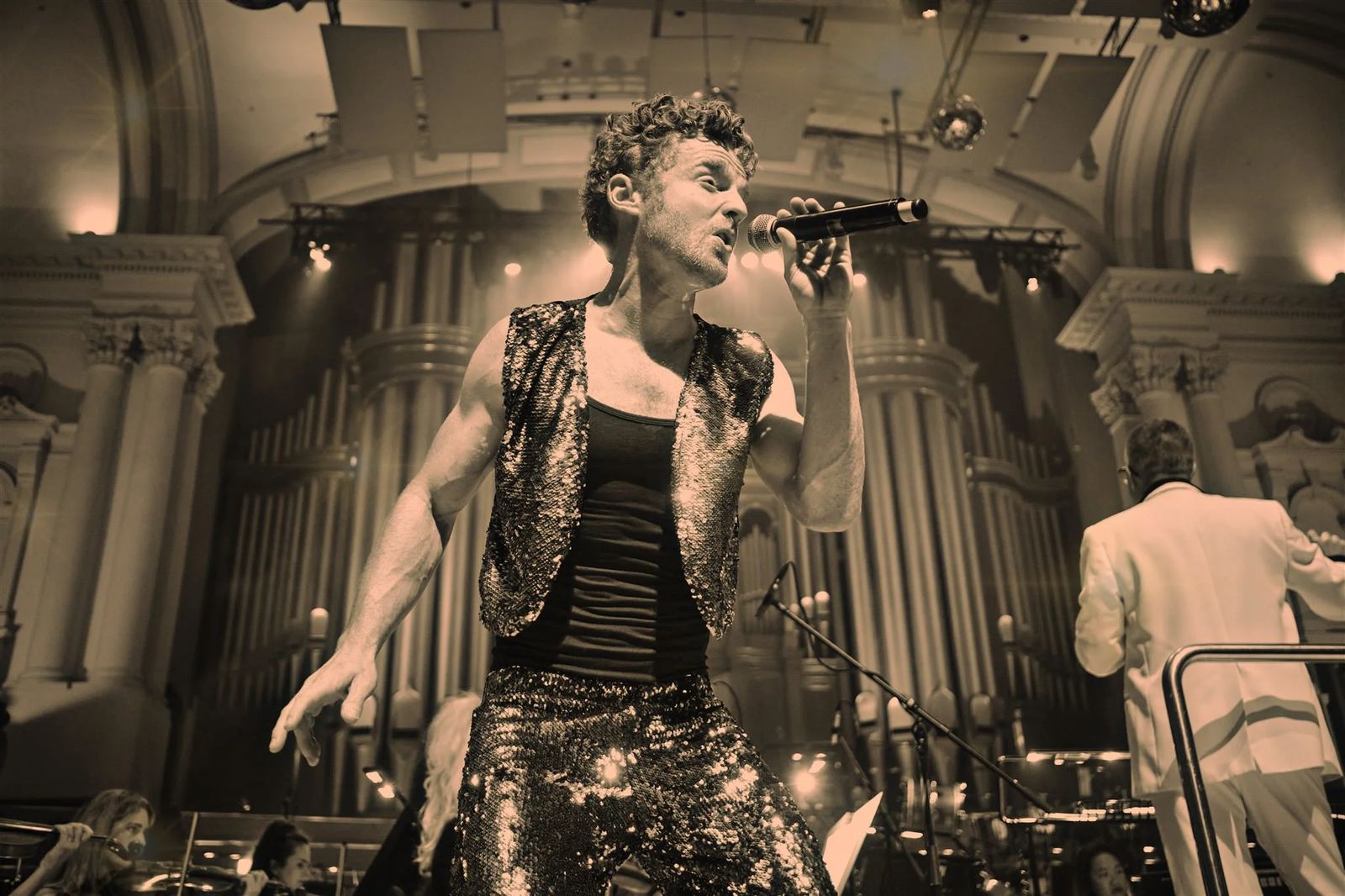 Dillon Rhodes singing under warm stage lights at Auckland Town Hall, wearing a sequinned vest with an orchestra behind him
