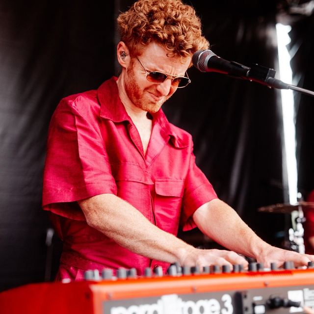 Dillon Rhodes playing keys at an outdoor festival in a red shirt and sunglasses