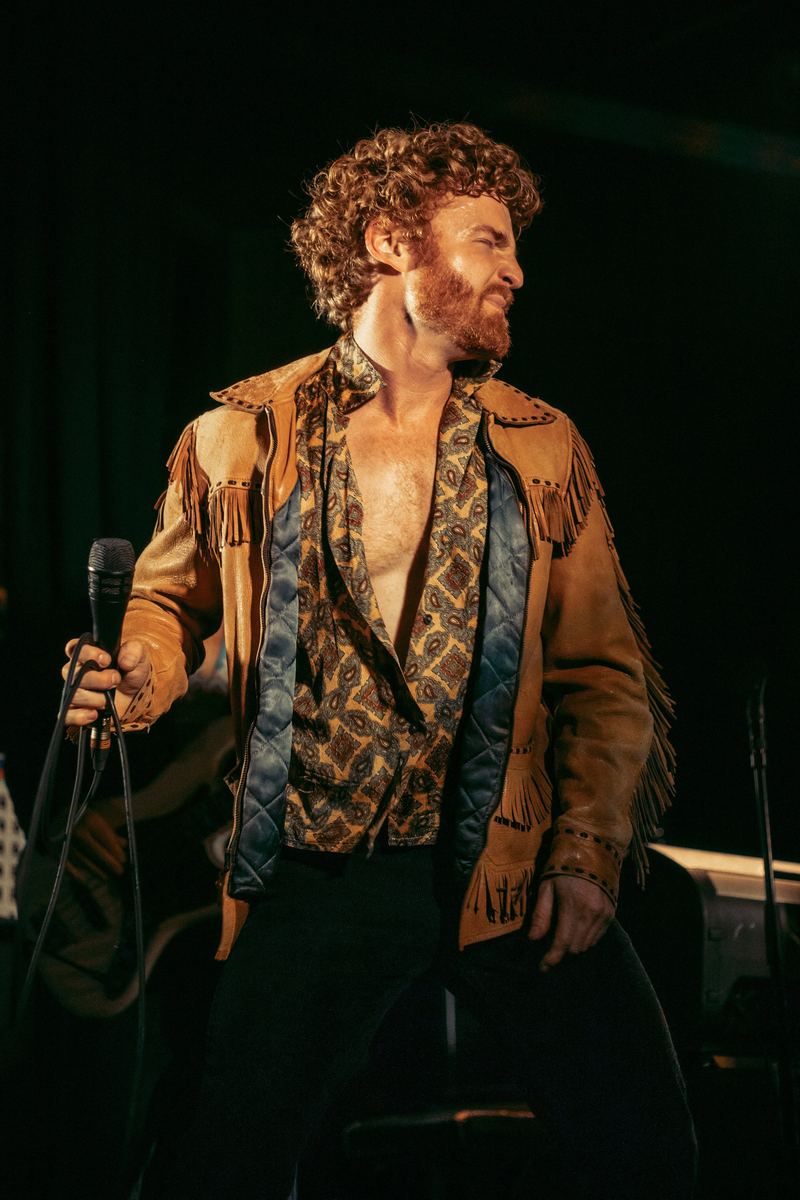 Portrait of Dillon Rhodes on stage in a fringed suede jacket and paisley shirt, holding a microphone in warm golden light
